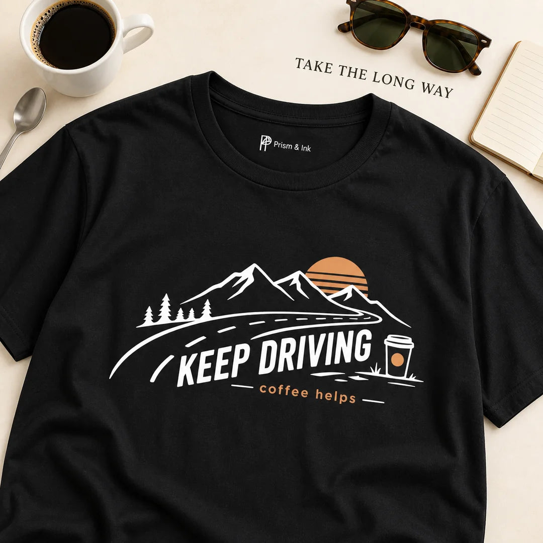 A black short-sleeve graphic T-shirt laid flat on a light beige surface with coffee, sunglasses, and an open notebook around it. The shirt has a scenic mountain and road illustration with the text 'KEEP DRIVING coffee helps' and a small 'Pious & Wise' brand mark printed inside the collar.